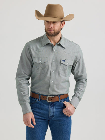 Man in cowboy hat, gray western shirt, and blue jeans standing against plain background