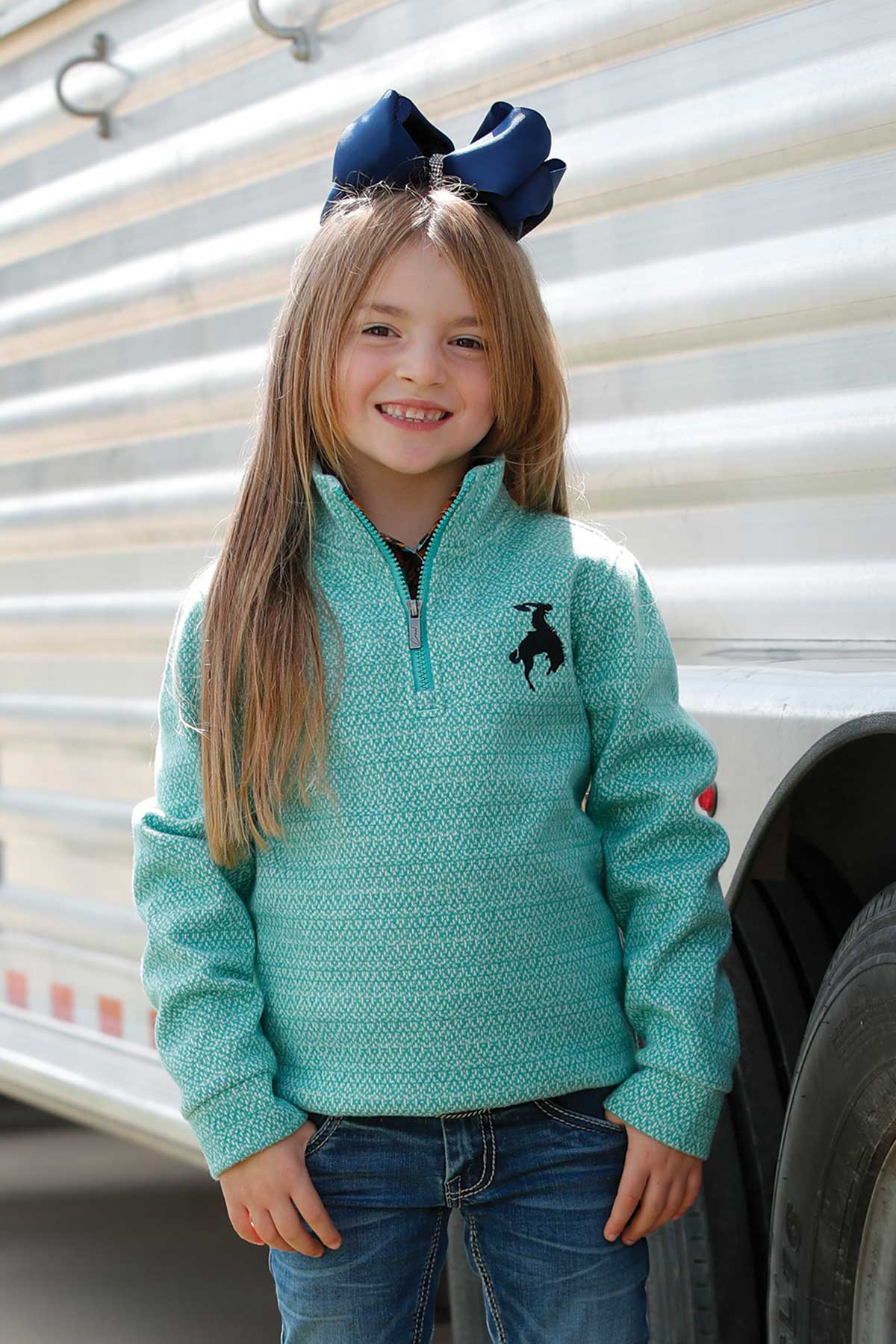 Smiling girl in turquoise quarter-zip pullover with black rodeo logo and blue bow, outdoors
