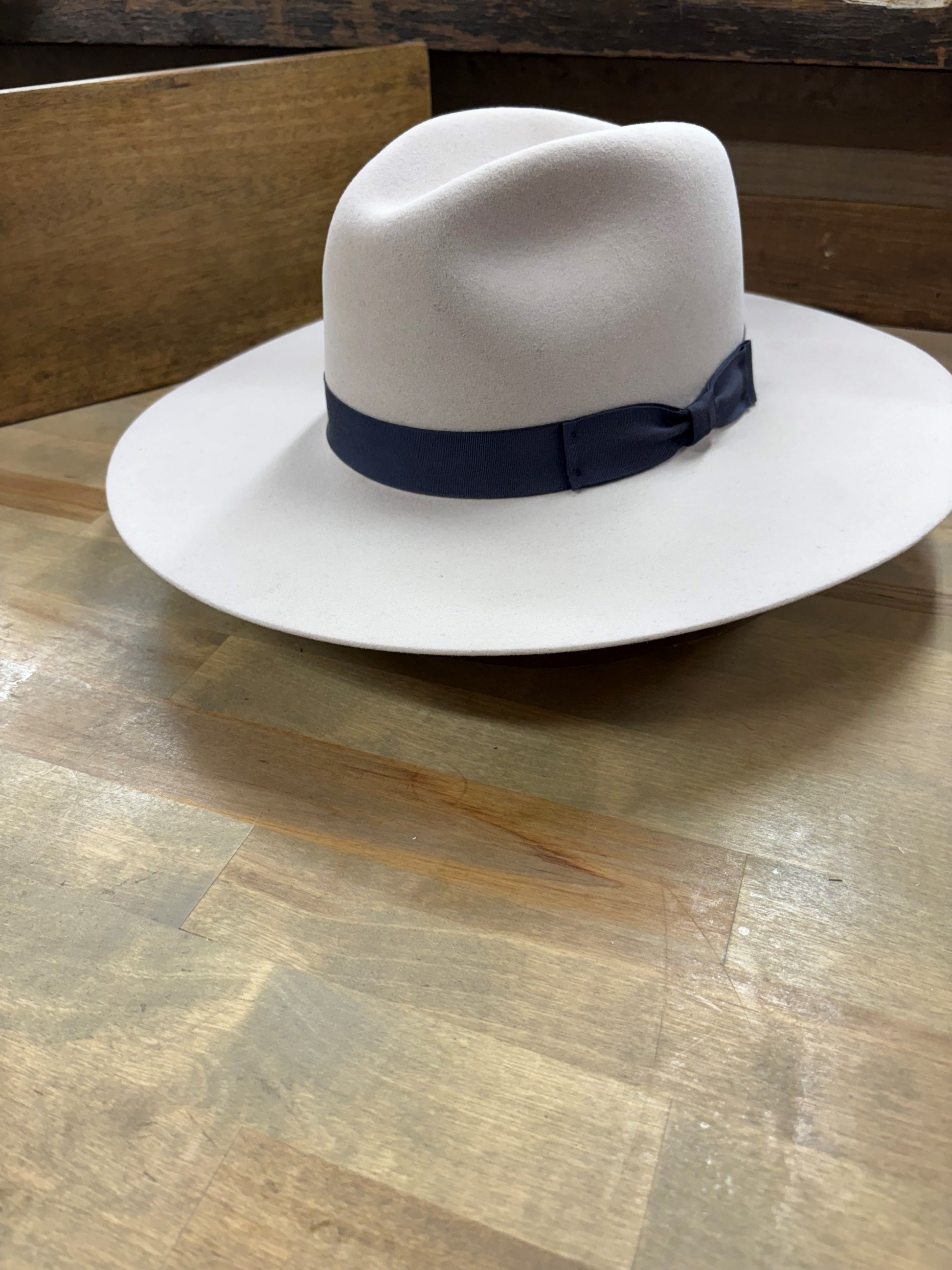 Light grey wide-brimmed fedora hat with a navy blue band on a wooden table.