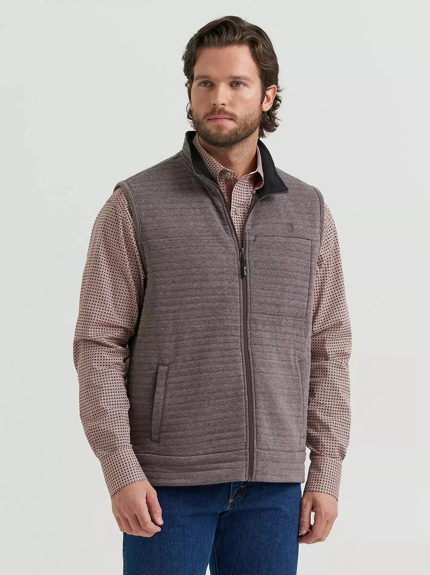 A man wearing a ribbed gray zip-up vest over a brown and white patterned long-sleeve shirt, paired with blue jeans, against a light gray studio background.