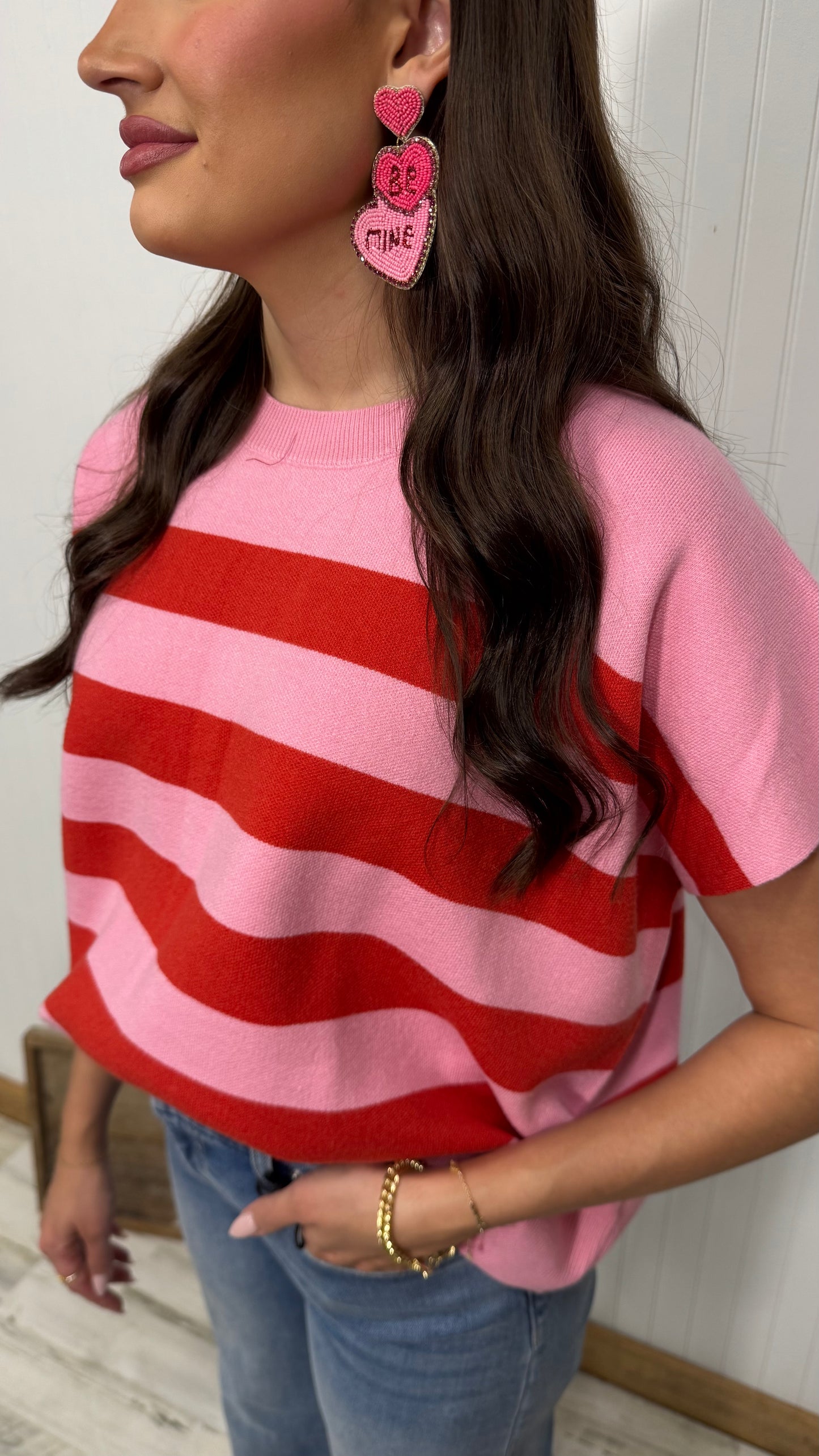 Woman in pink and red striped sweater, wearing statement heart earrings, blue jeans, gold bracelets.