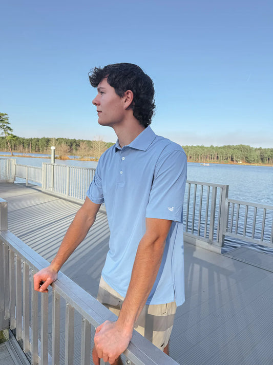 Person standing on a dock by a lake, wearing a light blue polo shirt.