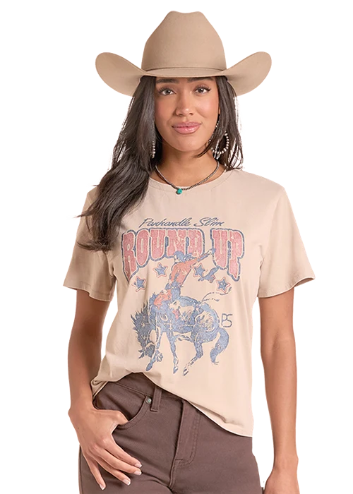 A woman wearing a tan cowboy hat and a beige graphic t-shirt with a blue and pink 'Round Up' western design, paired with brown pants, against a solid white background.