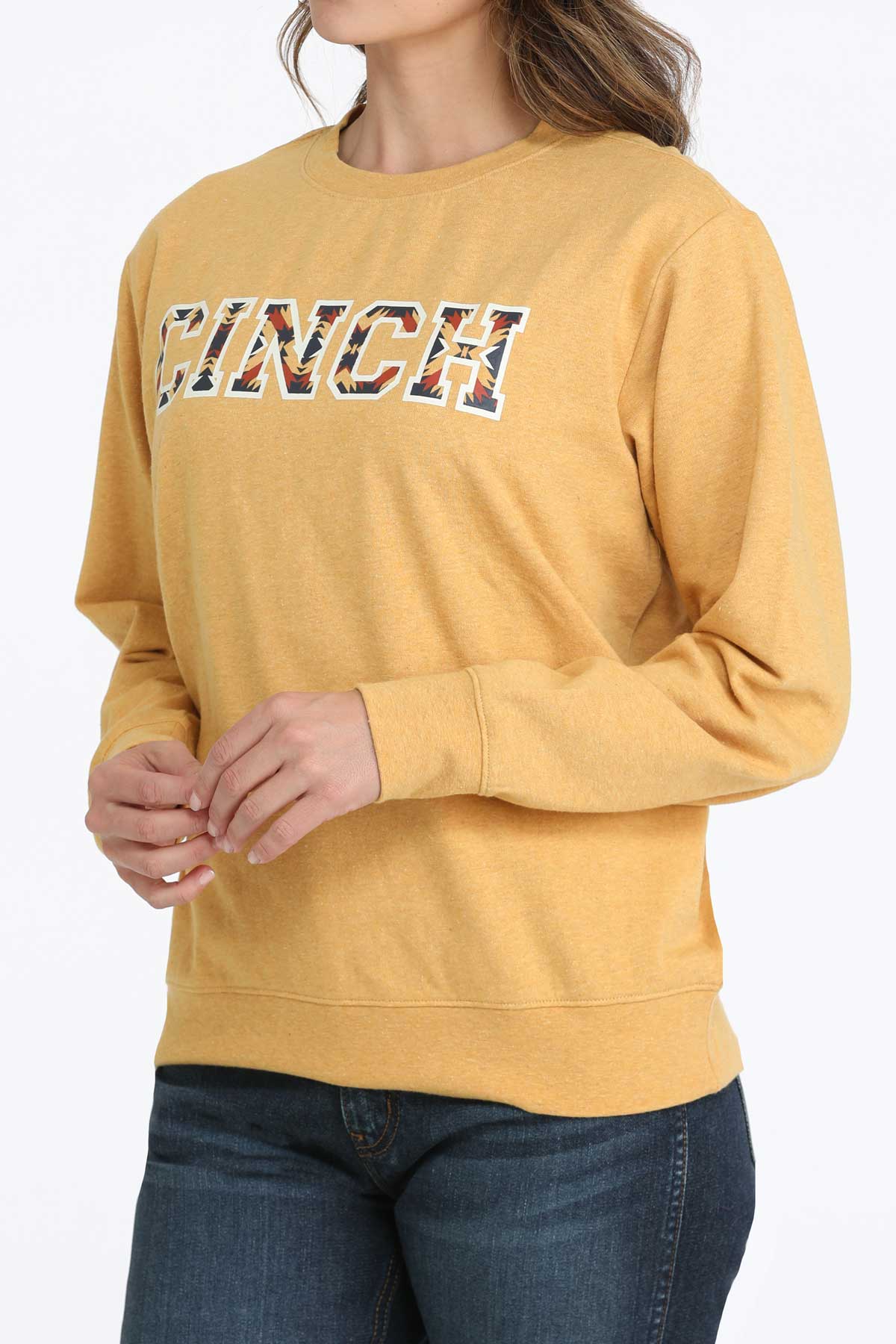 Woman wearing a mustard yellow CINCH graphic crewneck sweatshirt and dark jeans