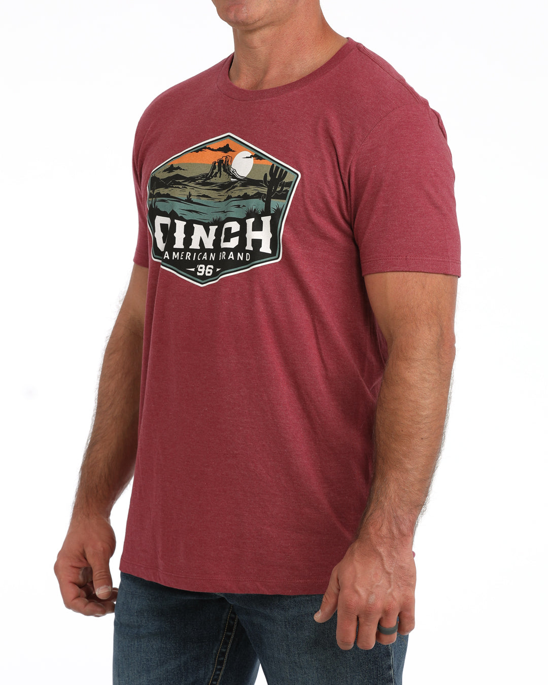 A man wearing a heathered red crewneck t-shirt with a large vintage-style graphic on the chest that reads 'CINCH American Flags 98' with a landscape illustration, paired with blue jeans against a white background.