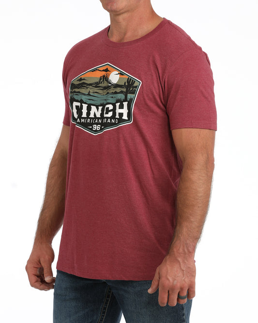 A man wearing a heathered red crewneck t-shirt with a large vintage-style graphic on the chest that reads 'CINCH American Flags 98' with a landscape illustration, paired with blue jeans against a white background.