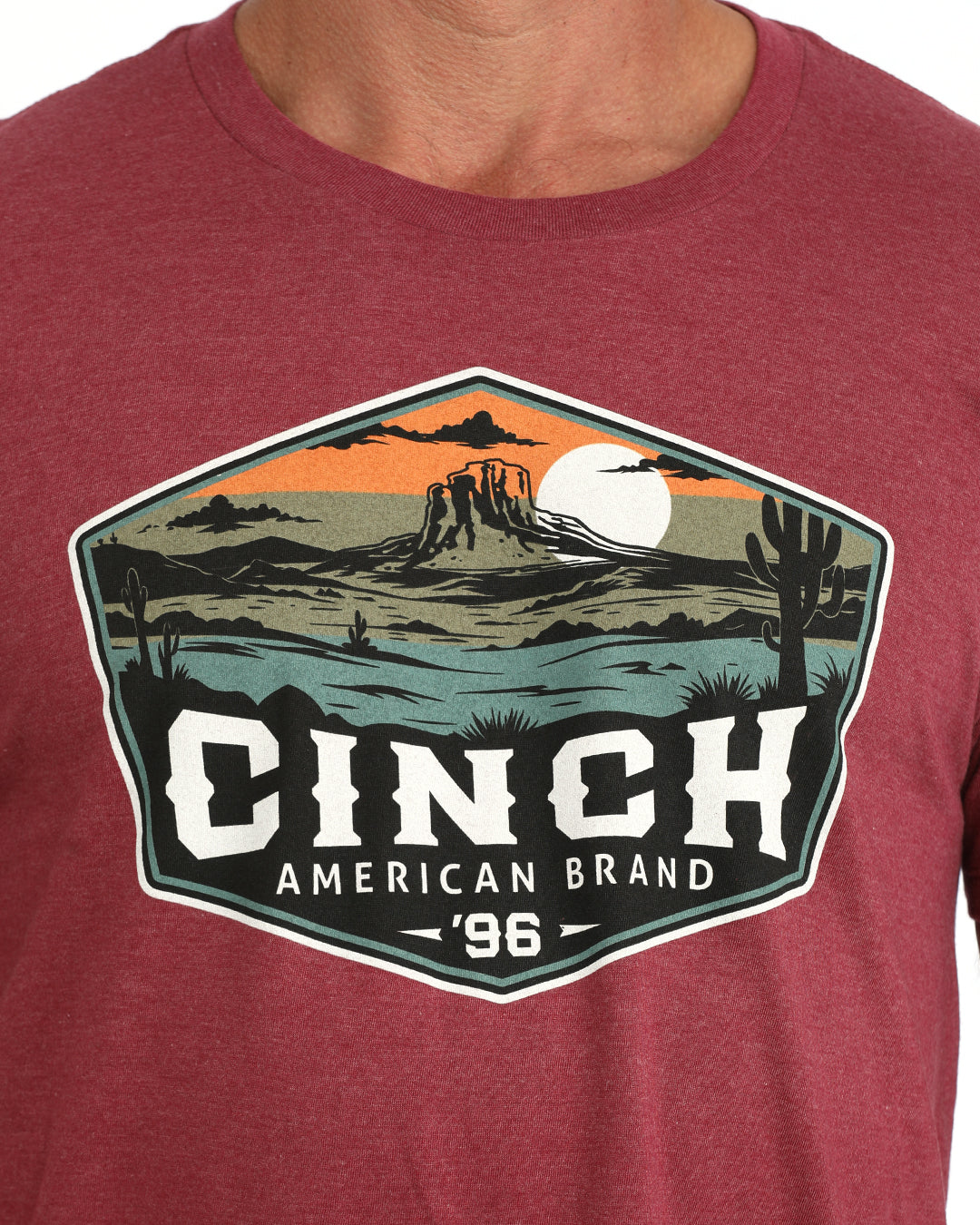 A close-up of a heathered red t-shirt featuring a large graphic print on the chest. The graphic depicts a desert landscape with a sunset, mountains, and cacti, with the text 'CINCH AMERICAN BRAND '96' printed below.