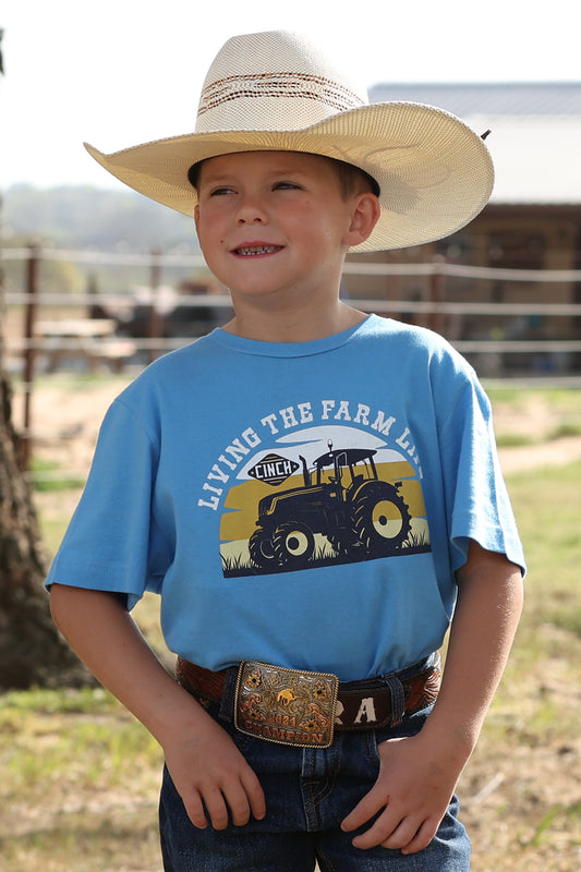 Child wearing a cowboy hat and blue t-shirt with a tractor graphic, standing outdoors.