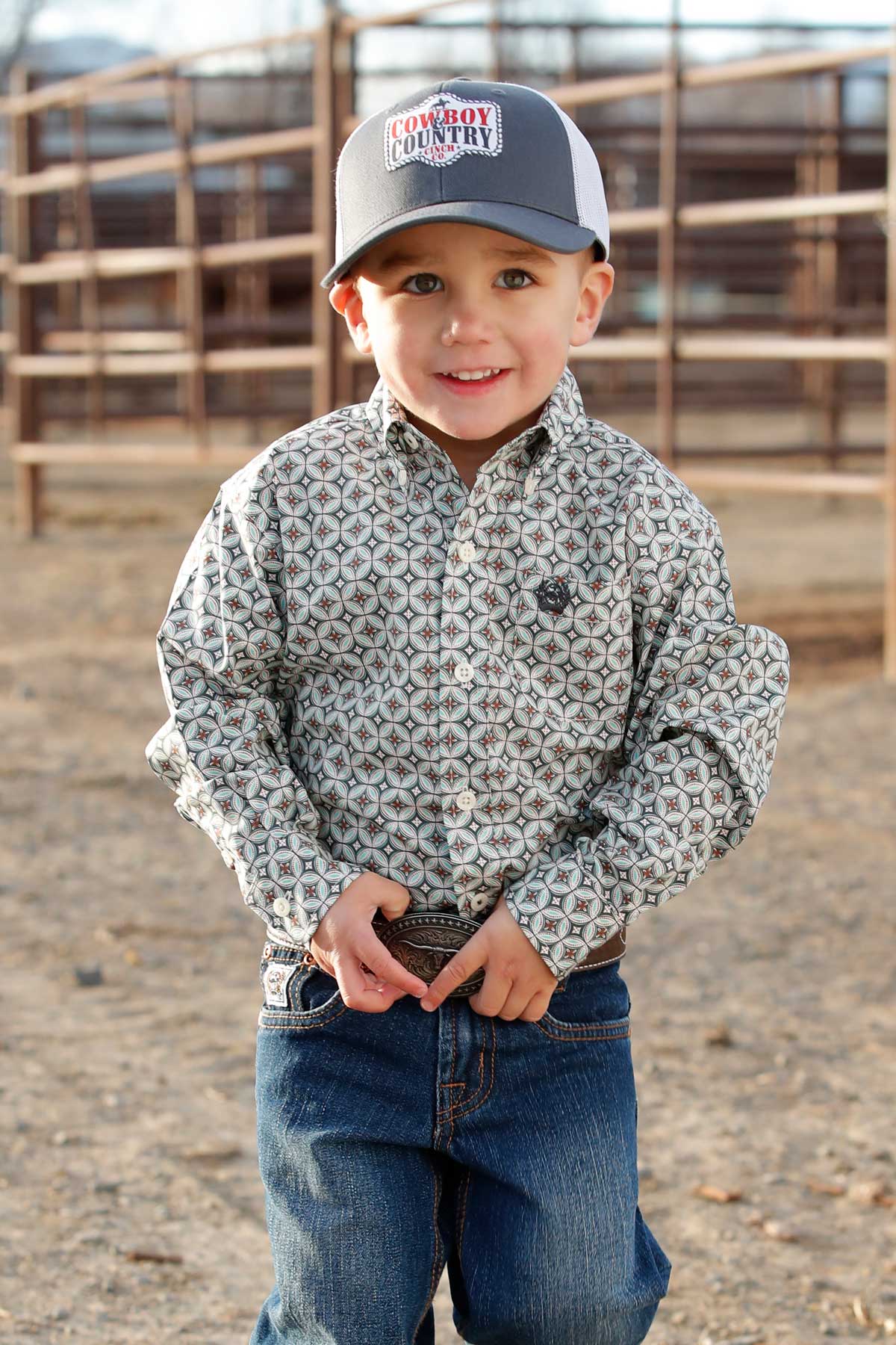 Smiling young boy in patterned western shirt, jeans, and cowboy hat at a ranch corral