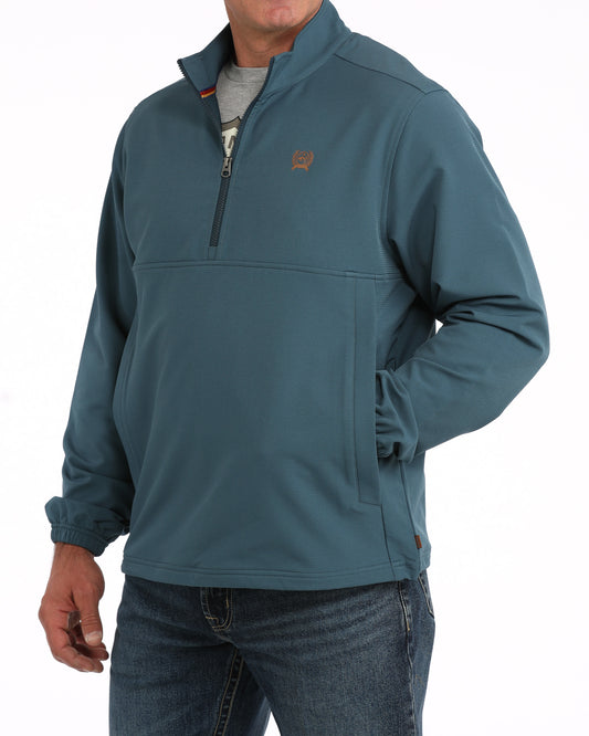 A man wearing a teal blue quarter-zip pullover jacket with a small brown embroidered logo on the left chest, paired with blue jeans.