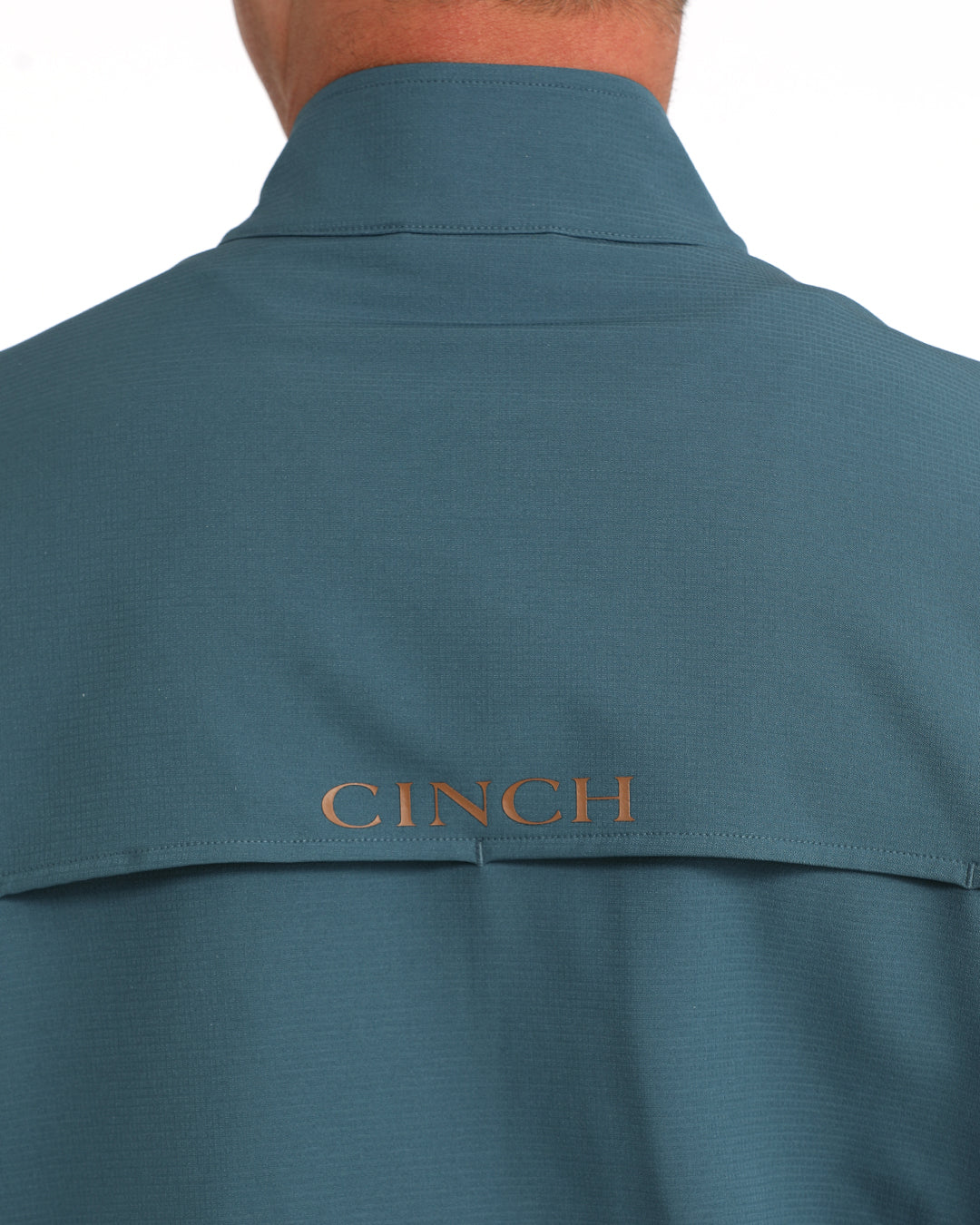 Close-up back view of a person wearing a teal-blue collared shirt with the brand name CINCH embroidered in gold on the upper back.