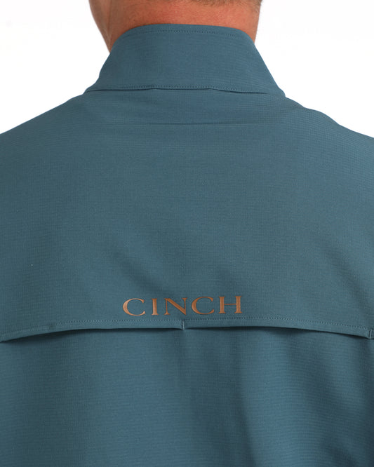 Close-up back view of a person wearing a teal-blue collared shirt with the brand name CINCH embroidered in gold on the upper back.