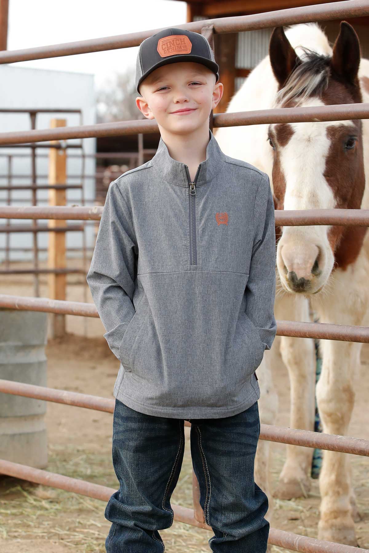 Smiling boy in gray quarter-zip jacket and cap standing by a horse at a ranch fence