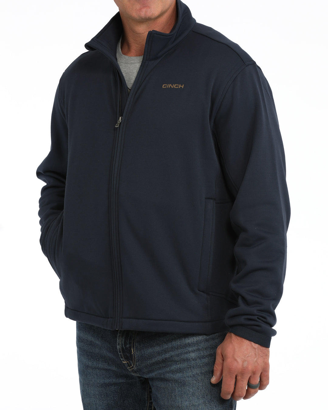 A man wearing a dark navy blue zip-up fleece jacket with a small gold 'PINEH' logo on the left chest, paired with a gray t-shirt and blue jeans, against a plain white background.