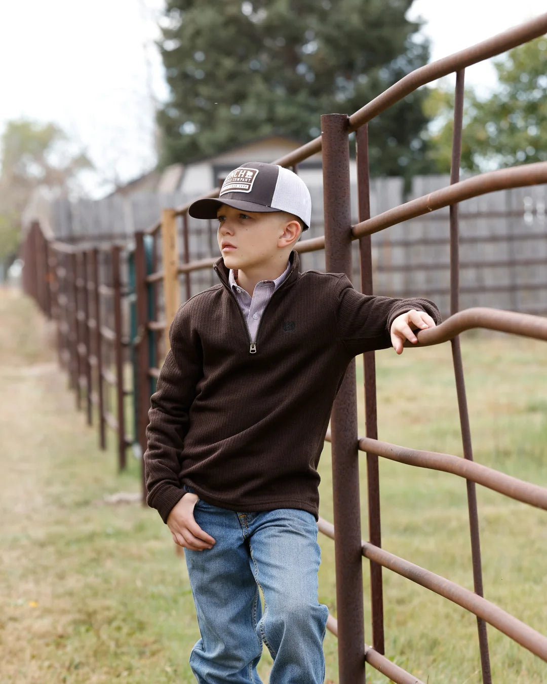 A young boy wearing a brown quarter-zip fleece pullover, blue jeans, and a black and white trucker hat leans against a rustic metal fence in an outdoor ranch setting.