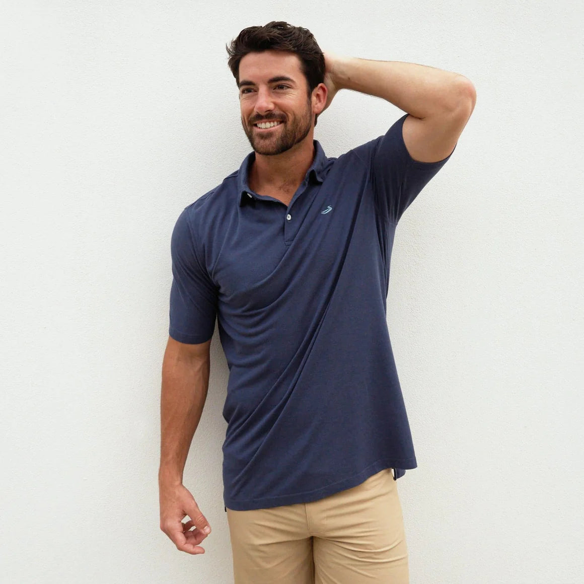 Man in navy blue polo shirt and khaki pants smiling against a plain white wall