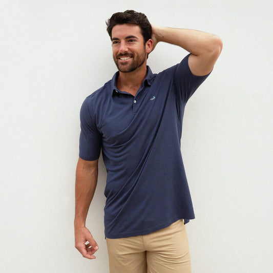 Man in navy blue polo shirt and khaki pants smiling against a plain white wall