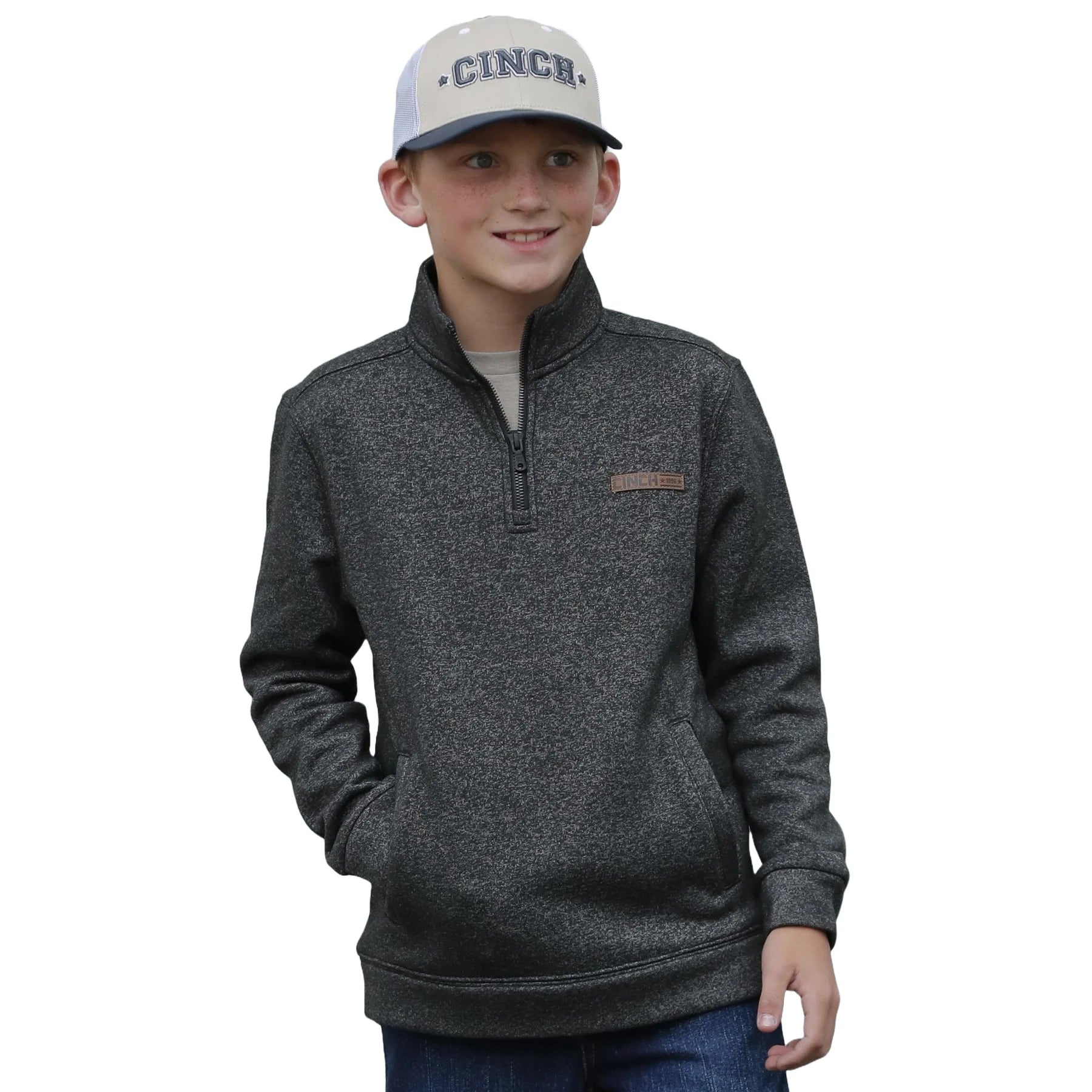 Boy wearing a grey quarter-zip pullover and blue trucker hat with hands in pockets