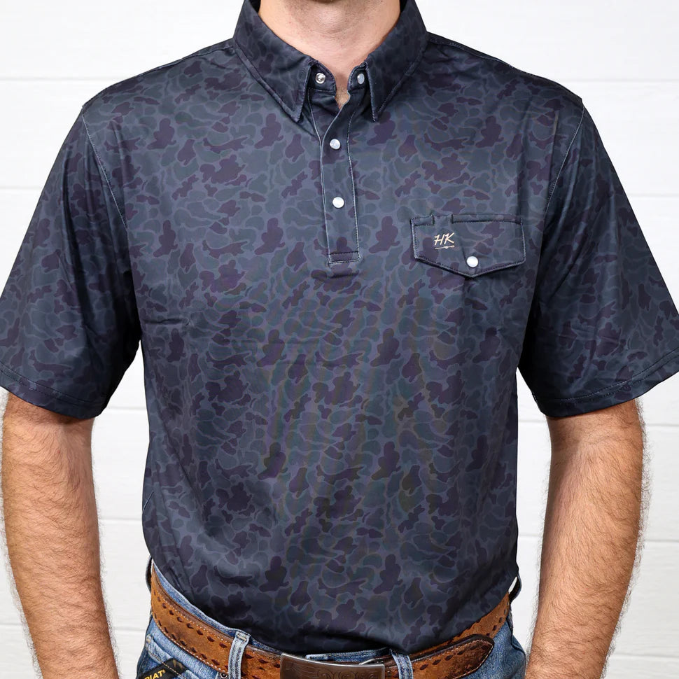Man wearing a dark patterned polo shirt against a white brick wall.