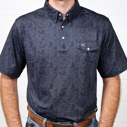 Man wearing a dark patterned polo shirt against a white brick wall.