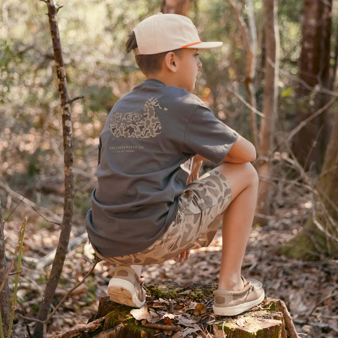 Boy in camo shorts and graphic t-shirt squatting on a tree stump in the forest