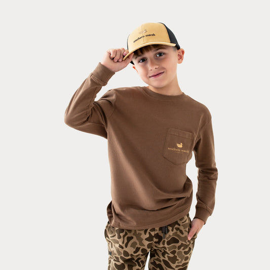 Boy wearing brown long sleeve shirt, khaki logo cap, and camo pants on white background