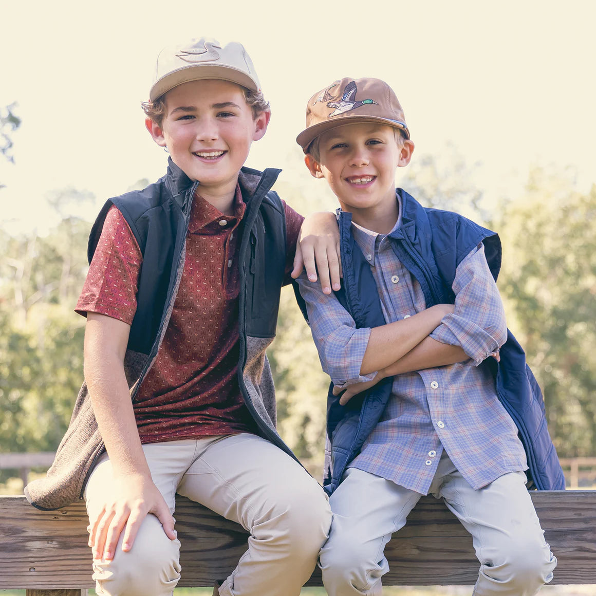 Two boys outdoors wearing casual shirts, vests, and hats, sitting on a wooden railing
