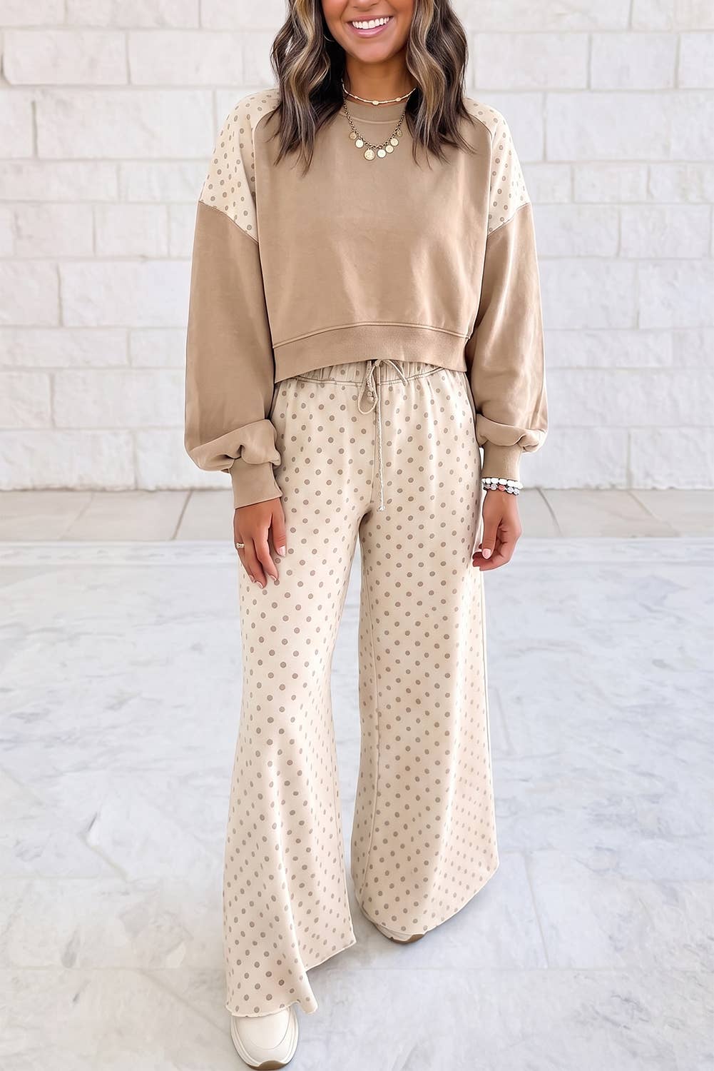 A woman modeling a beige cropped sweatshirt with sheer long sleeves and matching beige wide-leg pants with a small polka dot pattern, standing against a white brick wall.