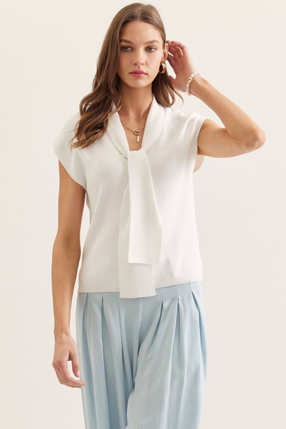 Woman wearing a white sleeveless blouse with a tie detail and light blue pants on a plain background