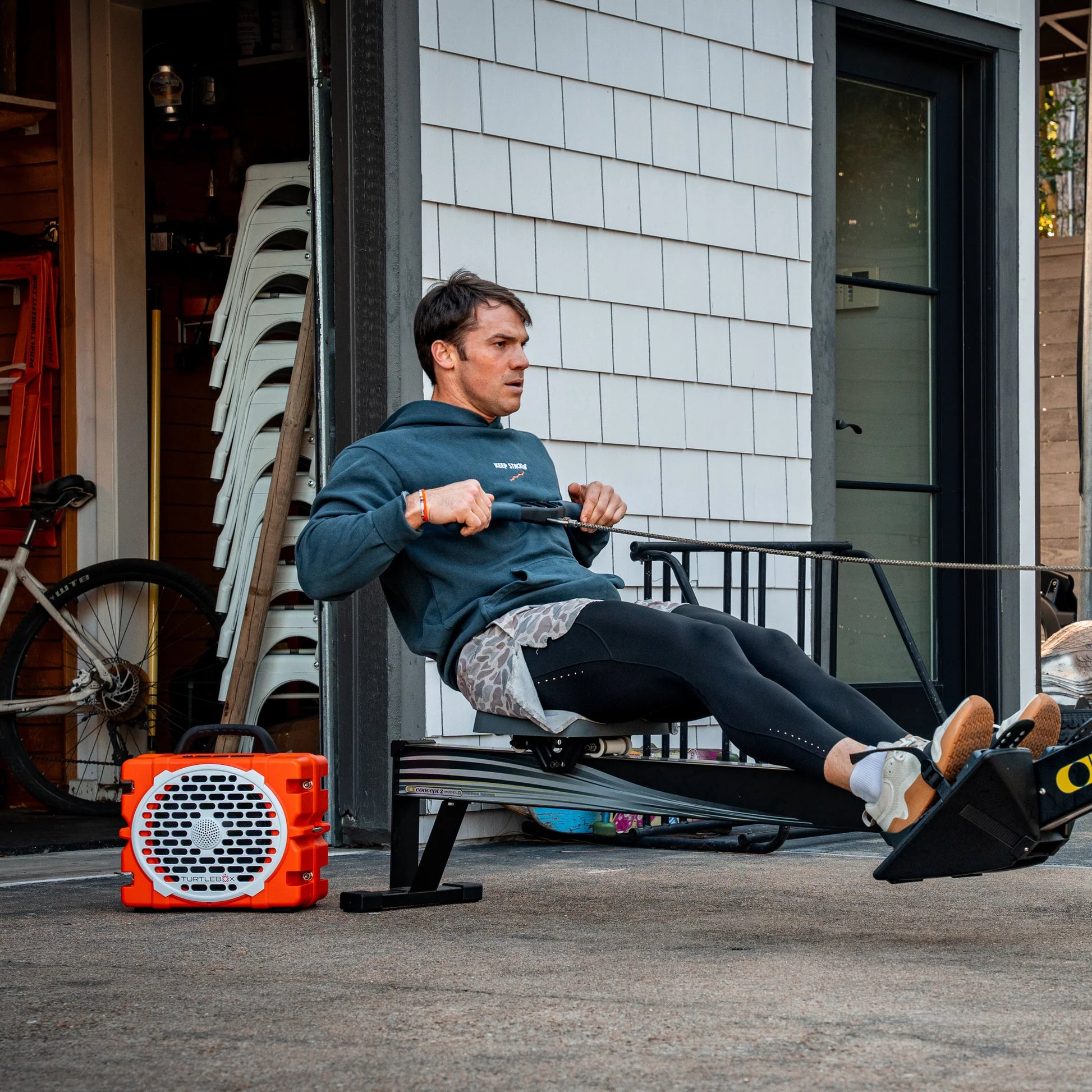 Man using a rowing machine outdoors with a building and trees in the background