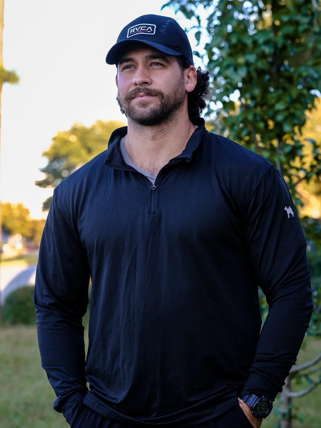 Man wearing a black jacket and cap with a logo, standing outdoors.
